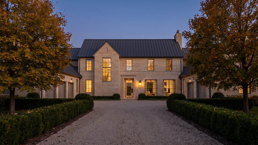 Limestone country house at the head of a gravel drive at dusk in early autumn, windows lit, maples turning gold behind it