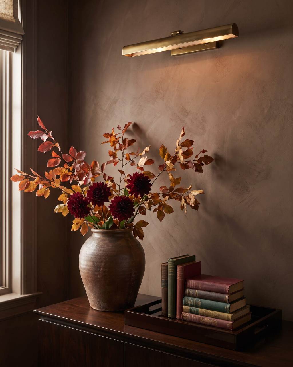 Hand-thrown earthenware vessel of turning branches and burgundy dahlias against a chocolate-brown plaster wall