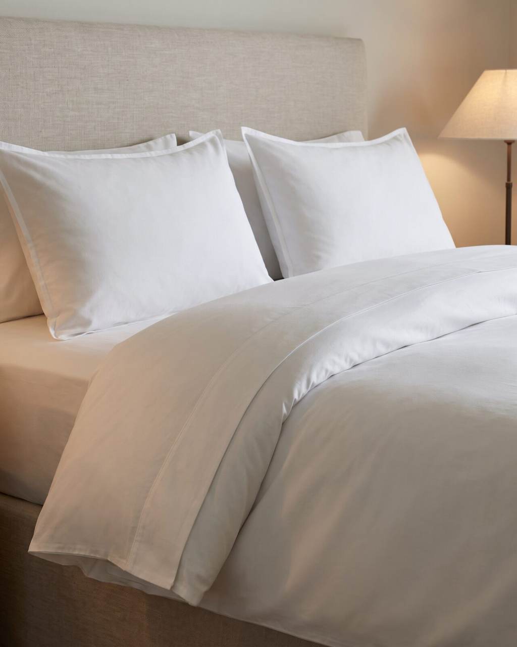 Close view of a guest bed dressed in matte white cotton percale, the flat sheet turned down over a lofty duvet, flanged pillowcases against an oatmeal linen headboard in warm lamplight
