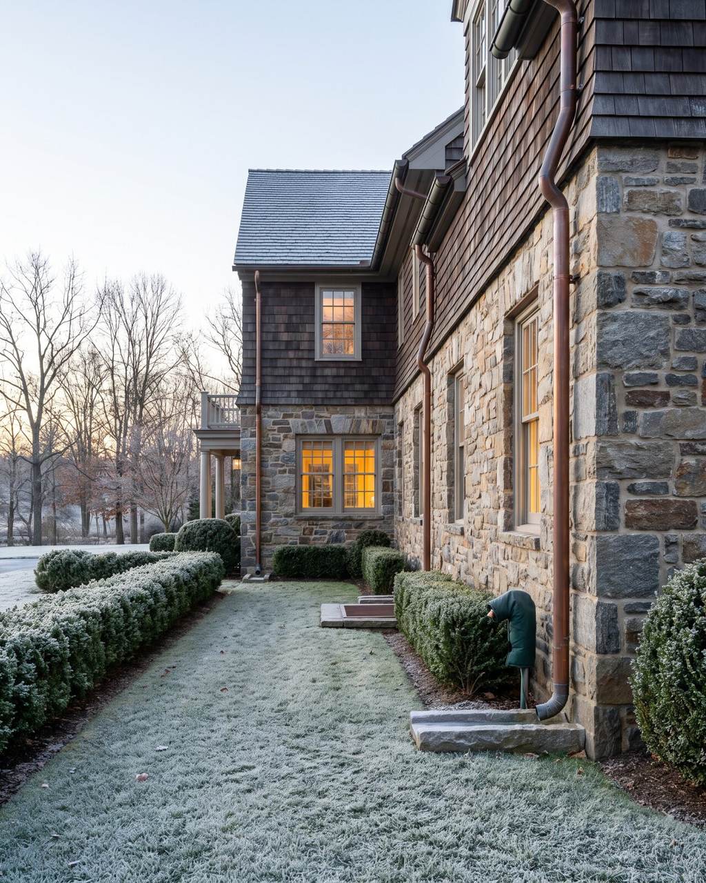 Side elevation of a stone-and-shingle house on a frosty morning, copper downspouts discharging onto stone splash blocks and an insulated cover fitted over the hose bibb