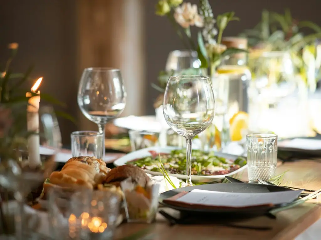 Candlelit wooden dining table set for dinner with wine glasses, dark plates, bread, a salad platter and low flowers