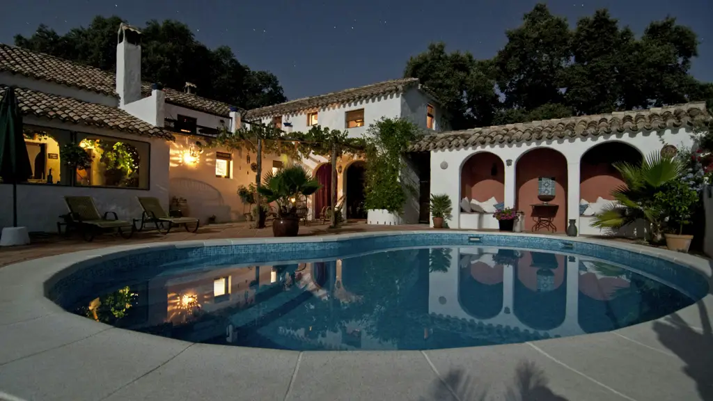 Whitewashed villa with a terracotta tile roof at night, lit windows and arched loggias reflected in a curved swimming pool under a starry sky