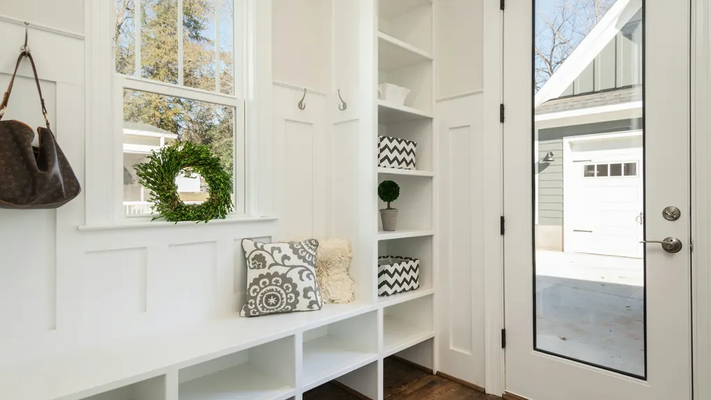 Bright white mudroom with a built-in bench, open cubbies, wall hooks and a glass door looking out to the garage