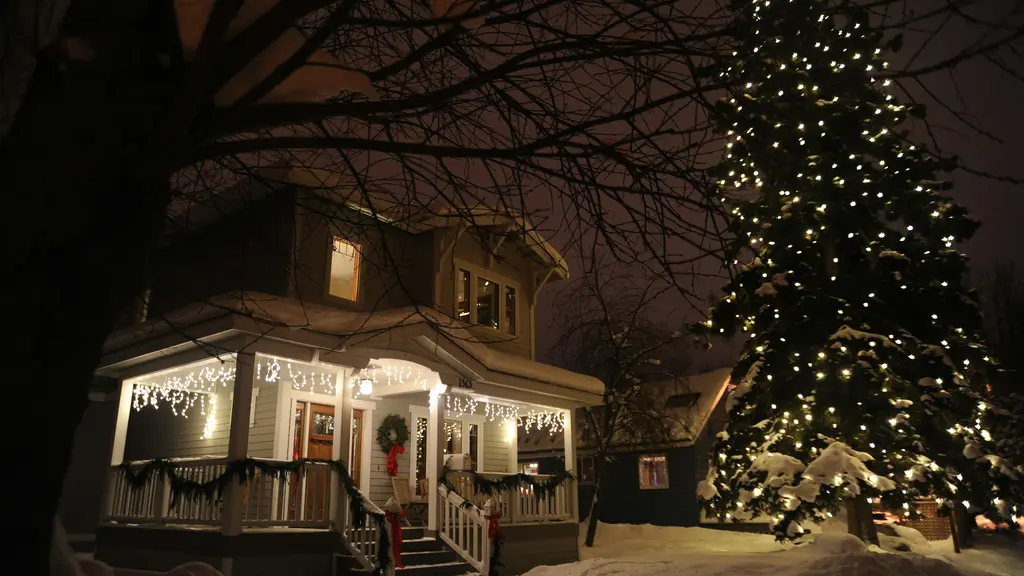 Two-story house at night in the snow, with warm white icicle lights along the porch roof, garland and a wreath at the front door, and a tall evergreen wrapped in white lights