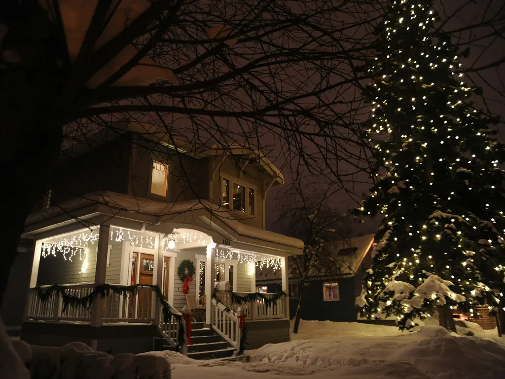 Two-story house at night in the snow, with warm white icicle lights along the porch roof, garland and a wreath at the front door, and a tall evergreen wrapped in white lights