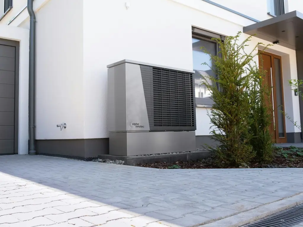 Grey outdoor mechanical unit on a gravel bed beside a white modern house, screened by young evergreen shrubs near the front door